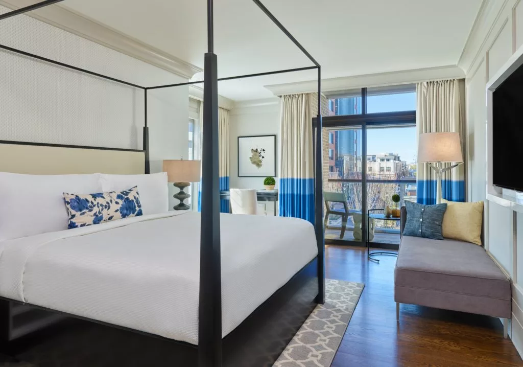 A photo of one of Hotel Avalon's Suite Bedrooms. The room is well lit with natural lighting streaming in through a balcony window. A large king-sized bed with white linens, a blue and white decorative pillow, and high bed posts that connect to an open canopy sits to the left of the photo. There's a couch with pillows on the right, sitting beneath a large screen television that hangs on the wall. Subtle accents of blue and white are throughout the room. There's a large rug underneath the bed which sits on dark wood floors. There's also artwork hanging on the wall.