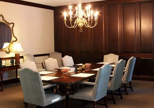 A photo of an elegant conference room with chandelier lighting. There are light blue/grey high-back chairs surrounding a long wooden table. There's also candy bowls on the table.
