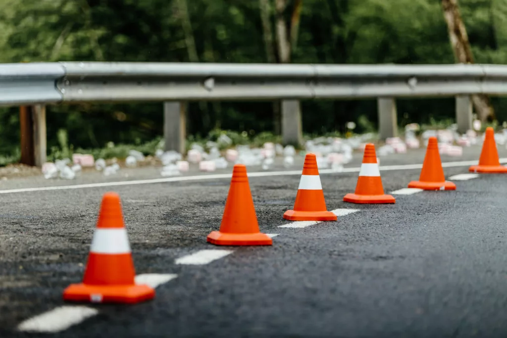 A close-up photo of a road or highway with orange cones lining it. There is glass and other debris next to a defective guardrail suggesting a car accident or crash of some kind has occurred.