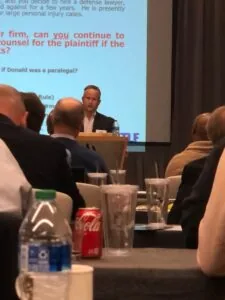 A photo of the backs of several people sitting in chairs with drinks. There is a man standing behind a podium in front of them with a large screen behind him.